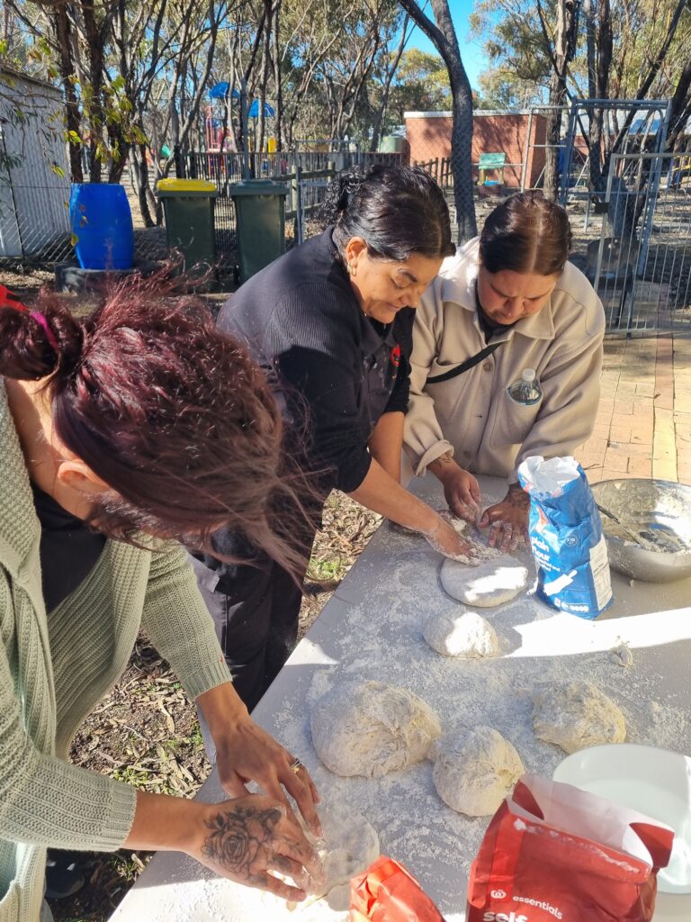 Three women standing around an outdoor table kneading some damper mixture.