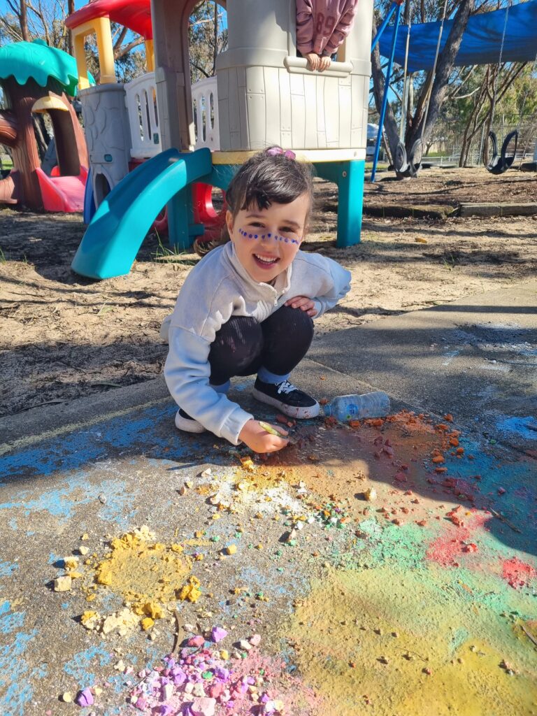 Child squatting as she is painting some concrete with water and smashed up pieces of coloured chalk on the ground in front of her, with colour smudged across the ground.