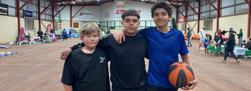 3 boys standing in the foreground with arms around each other and one holding a basketball. There's a stadium in the background and people with stalls set up around the perimeter.