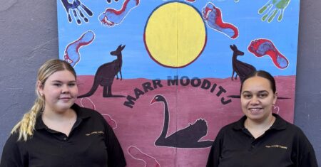 Two women facing the camera while they stand in front of a painting that includes the word Maar Mooditj and depicts a black swan, kangaroos, sun and foot and handprints.