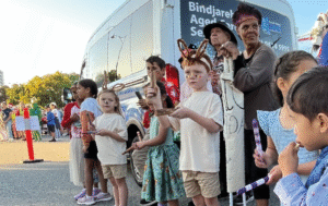 Preschool aged children playing clapping sticks while they stand alongside a road with two Aboriginal Elders standing behind them.