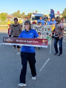 Woman holding a Bindjareb Aged Care banner and walking on a road with others walking behind them also holding a larger handpainted banner saying the same thing.