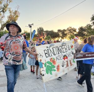 Aboriginal Elder man holding one side of a Bindjareb Aged Care banner while children and adults walk behind them. The sun is setting in the sky.