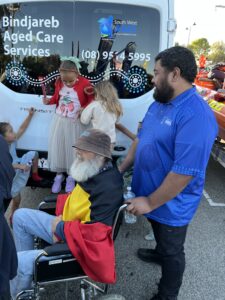 Man in a blue uniform holding the arms of a wheelchair and smiling at children. A man in a wheelchair has an Aboriginal flag draped over his upper body.