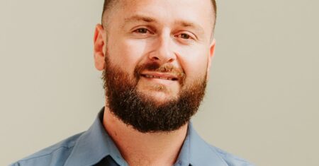Man with a beard and wearing a blue shirt with a logo. He is looking at the camera.